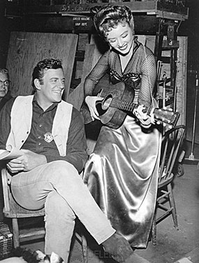 Taking a break from filming “Gunsmoke”, James Arness smiles as Amanda Blake 
fiddles with a gee-tar.