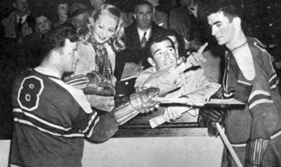 Adele gets instructions on how to hold a hockey stick from player Paul Courteah while Rod chats with Joe Kaholec.