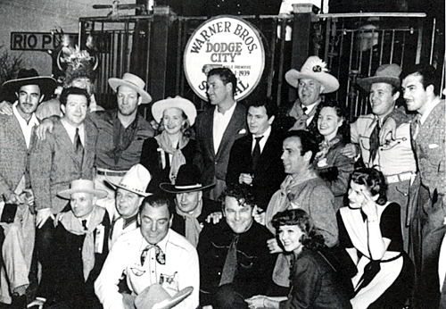 On tour in New Mexico to promote Errol Flynn’s “Dodge City” are (L-R) Gilbert Roland, Frank McHugh, Chief Santa Fe (behind McHugh), Maxie Rosenbloom, Priscilla Lane, Errol Flynn, John Garfield, Jack L. Warner (with hat on), Rosemary Lane, Wayne Morris and John Payne. (Kneeling) Leon G. Turrow, Hoot Gibson, Buck Jones, Lee Lyles (assistant to the President of the Santa Fe Railroad), Guinn "Big Boy" Williams, Humphrey Bogart, Jean Parker and Frances Robinson. Ten and fifteen minute stops were arranged for San Bernardino, Barstow, Needles, Seligman, Winslow, Gallup, Albuquerque, La Junta, Syracuse, NY and Chicago. Then back to Dodge City.