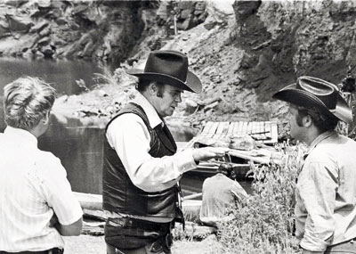 Sunset Carson and crew members prepare for the blowing up of the hideout shack scene from “Marshal of Windy Hollow” in the two photos above. In the first photo are Sunset, cameraman Darrell Cathcart and Jerry Whittington. In the second photo are an unknown lady, Sunset and Jerry Whittington.